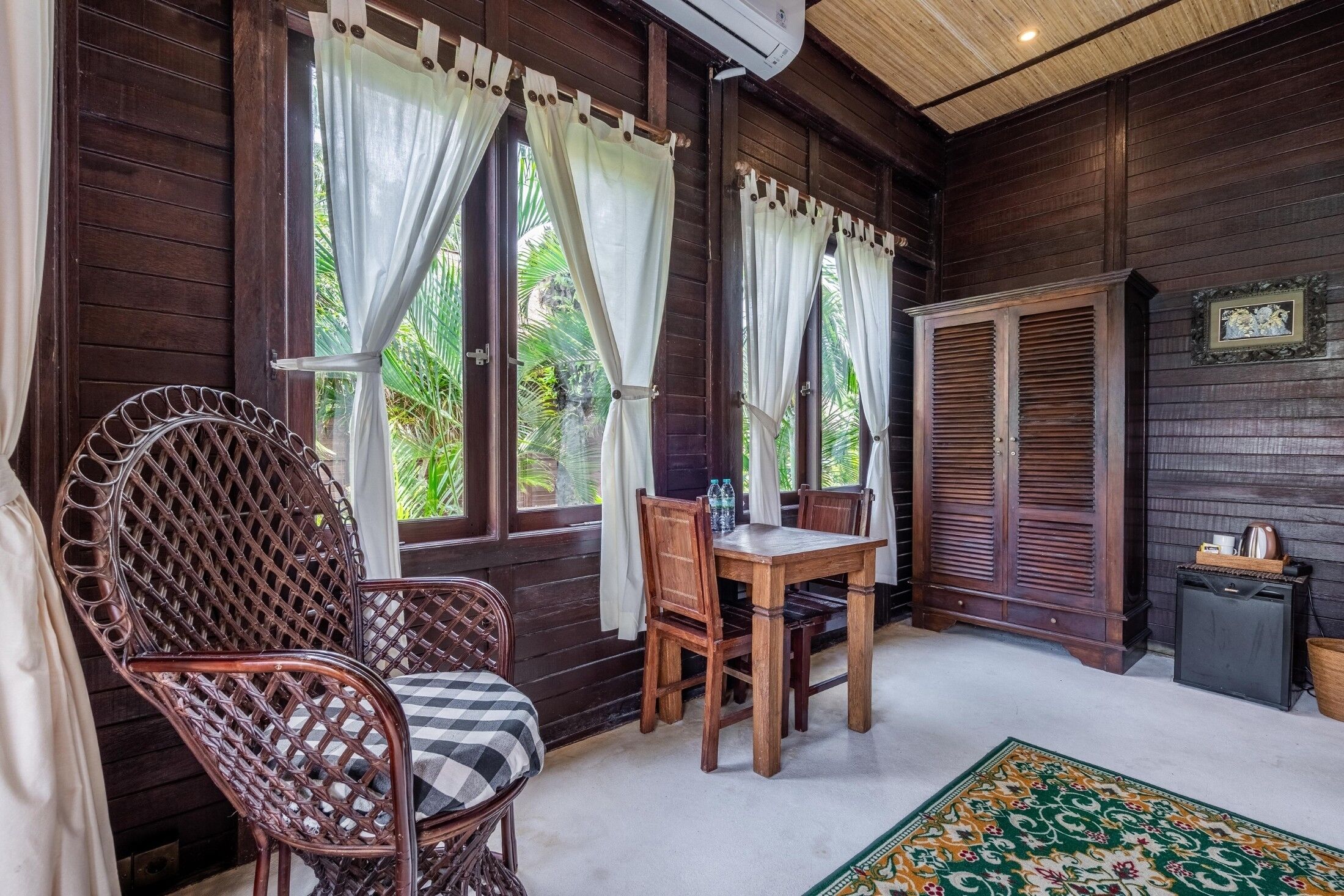 Una habitación de madera con una mesa y sillas, ventanas amplias con cortinas y una silla de ratán tradicional.