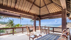 Een overdekt buitengebied met een bed, stoelen en een tafel. Er is uitzicht op de oceaan en palmbomen.