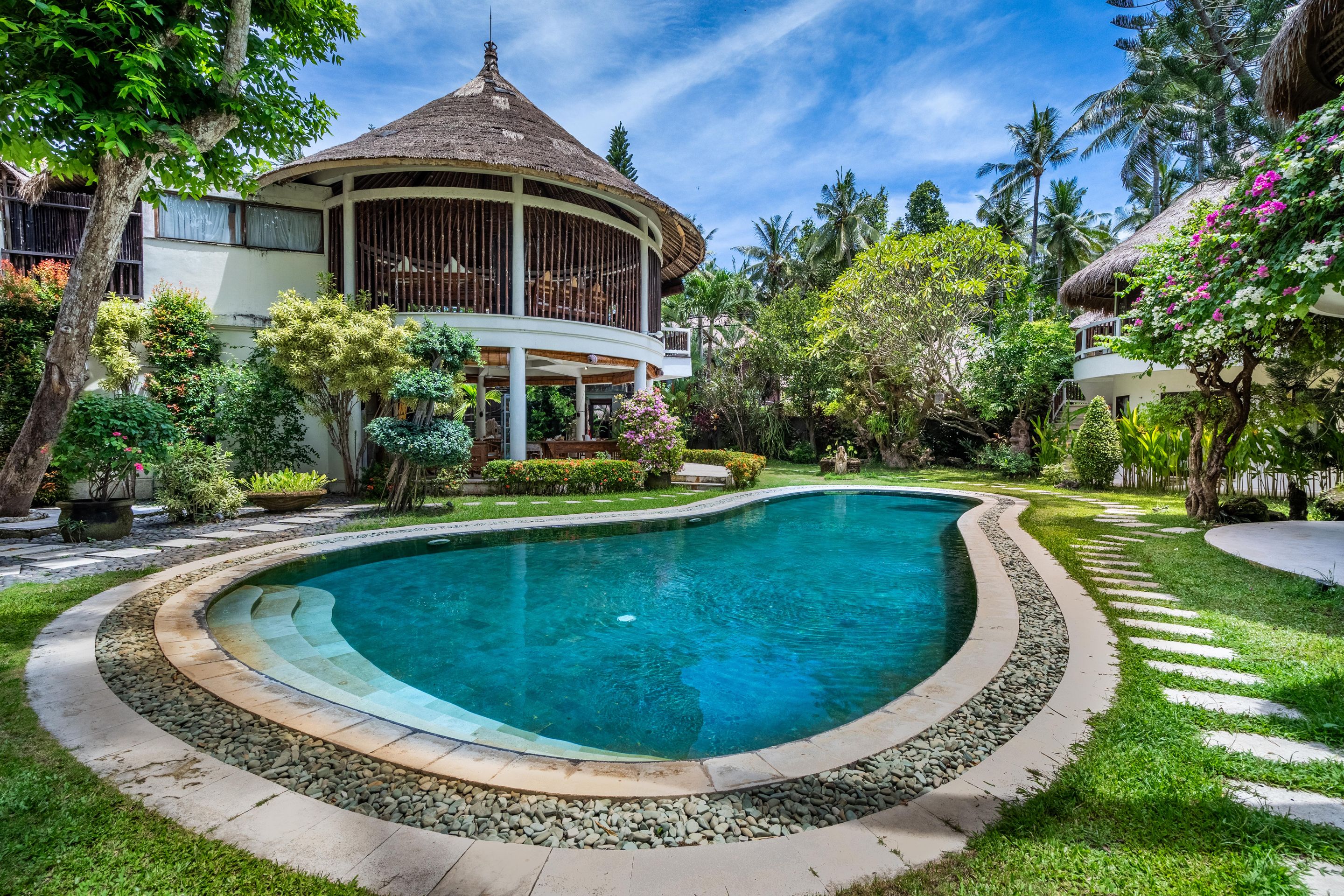 A circular swimming pool with a stone border, surrounded by lush greenery and a thatched-roof building.