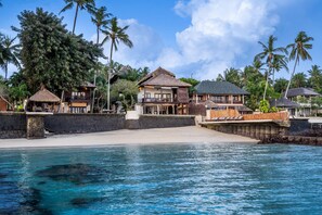 Een strandresort met meerdere bungalows boven het water, een zandstrand en helderblauw water.