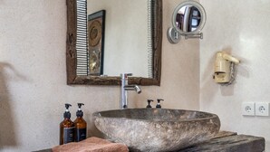 Een badkamer met een ruime stenen wasbak, een houten wastafelmeubel, een spiegel en twee flessen shampoo.