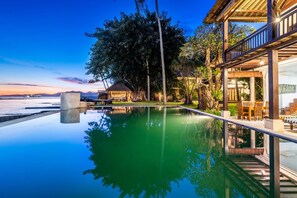 A serene pool area with a clear view of the sunset over the water, surrounded by lush greenery and a wooden deck. - Superior Oceanview Group Stay – 2 Levels, Separate Entrances | The Village of Angels Resort (Kabupaten Karangasem)