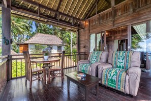 A wooden deck with a dining table and chairs, a sofa with a patterned throw, and a view of a house with a thatched roof. - Superior Oceanview Group Stay – 2 Levels, Separate Entrances (Kabupaten Karangasem)