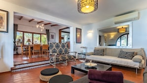 A living room with a sofa, coffee table, patterned armchair, dining area with chairs, and a kitchen in the background.