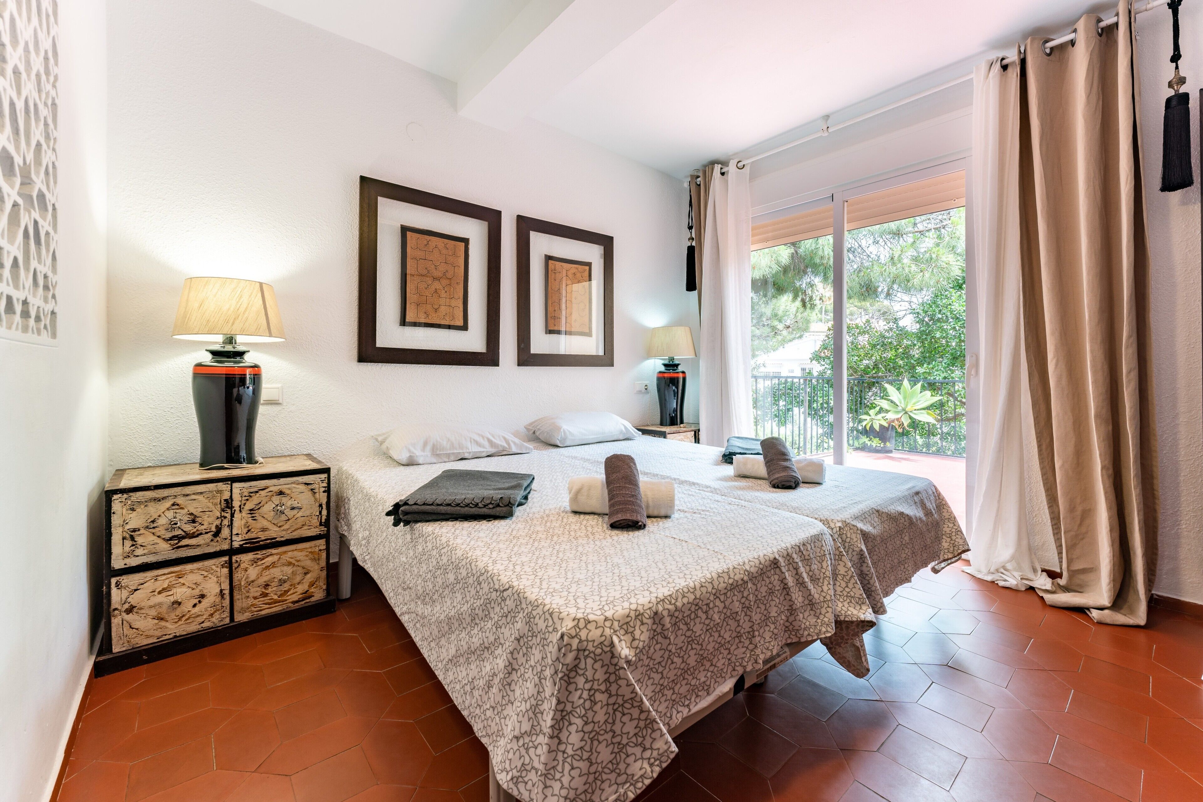 A hotel room with two beds, a nightstand with a lamp, a dresser, and a large window with curtains.