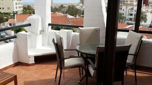 Un balcon avec une table ronde en verre et quatre chaises, des piliers blancs et une vue sur des immeubles et des palmiers.