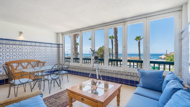 Een balkon met uitzicht op zee, een houten tafel, blauwe banken en betegelde vloeren.