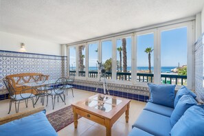 Een balkon met uitzicht op zee, een houten tafel, blauwe banken en betegelde vloeren.