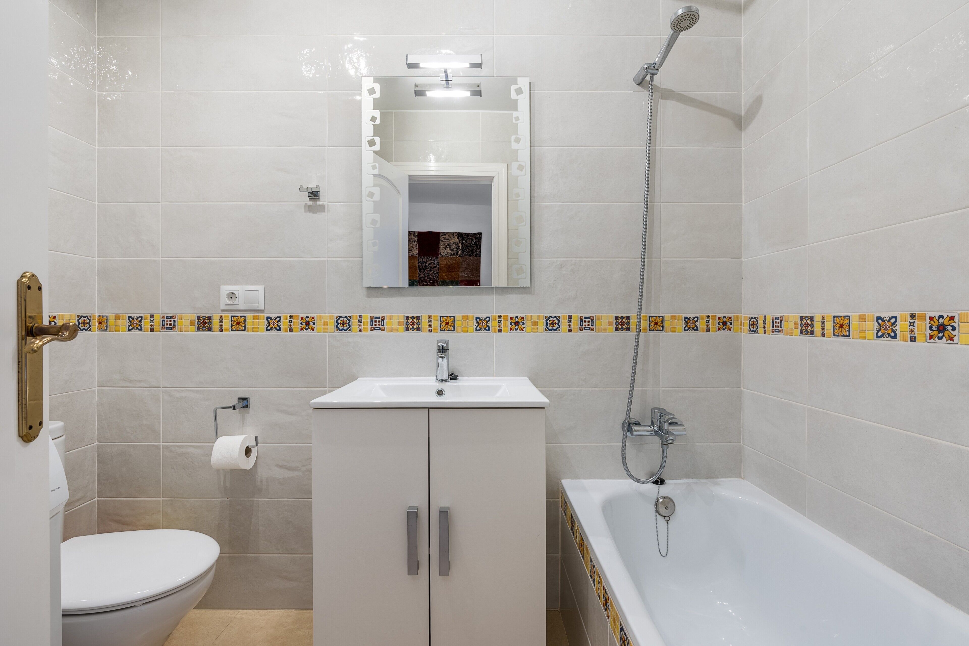 Casa de banho com sanita, lavatório, espelho, chuveiro e moldura de azulejos decorativa.