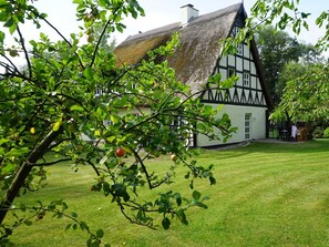 Ein traditionelles Fachwerkhäuschen mit Strohdach, umgeben von einem gepflegten Rasen und Obstbäumen.