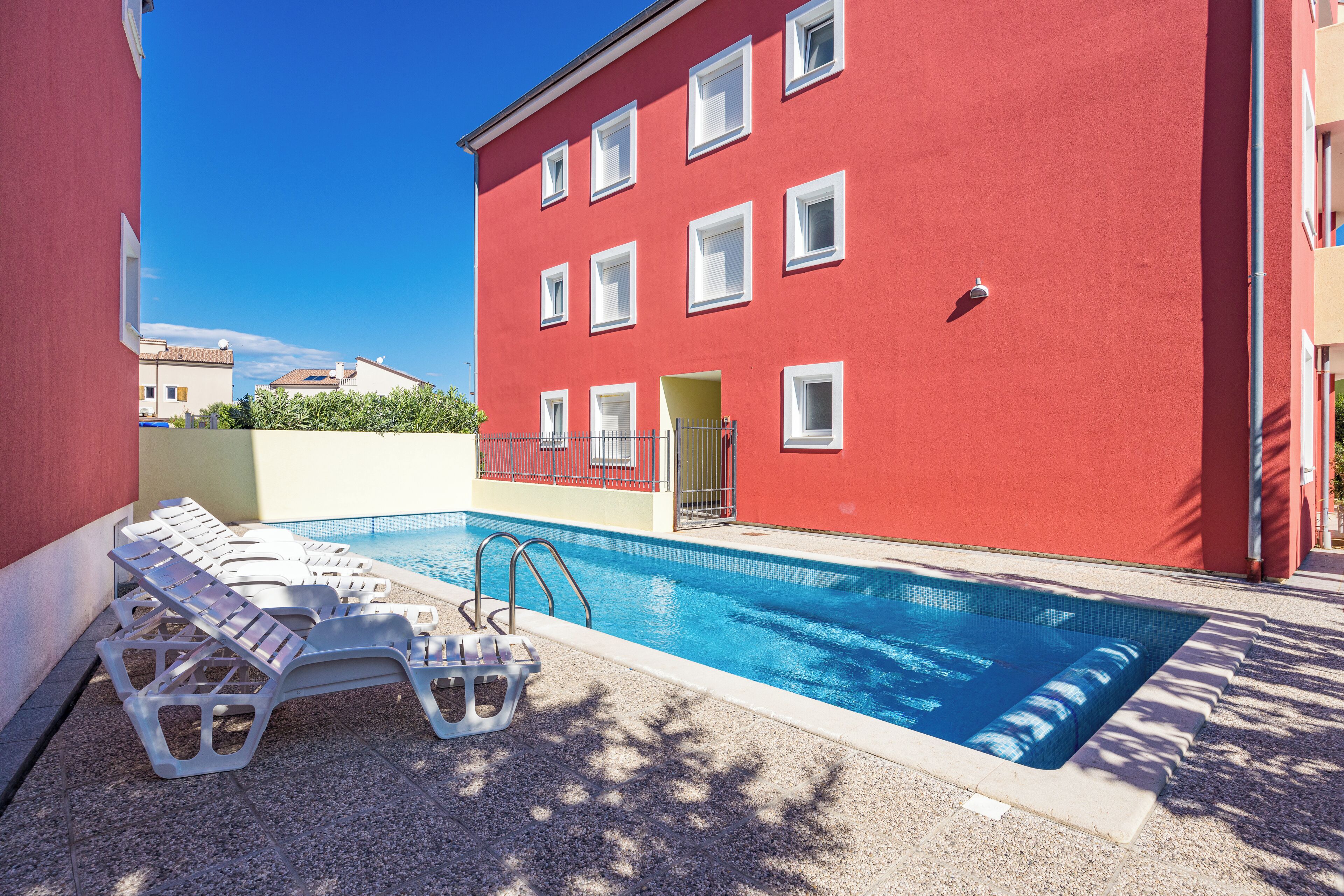 A red building with a swimming pool, lounge chairs, and a clear blue sky.