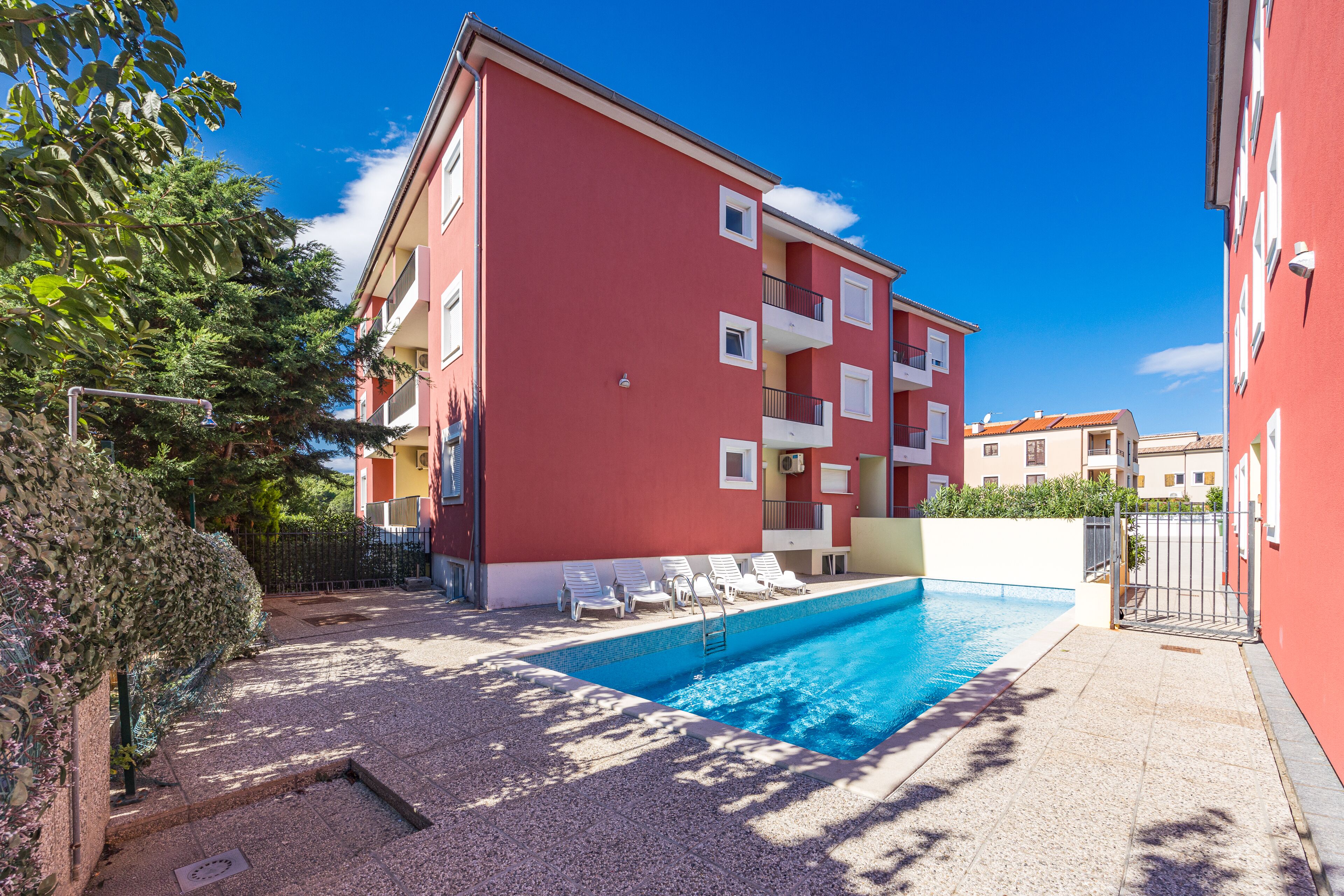 A red building with a swimming pool, white balconies, and a gated entrance.