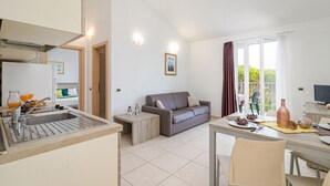 A modern kitchen with a sink, a dining area with a table and chairs, a sofa, and a large window with curtains. - Residence Kamenjak (Medulin)