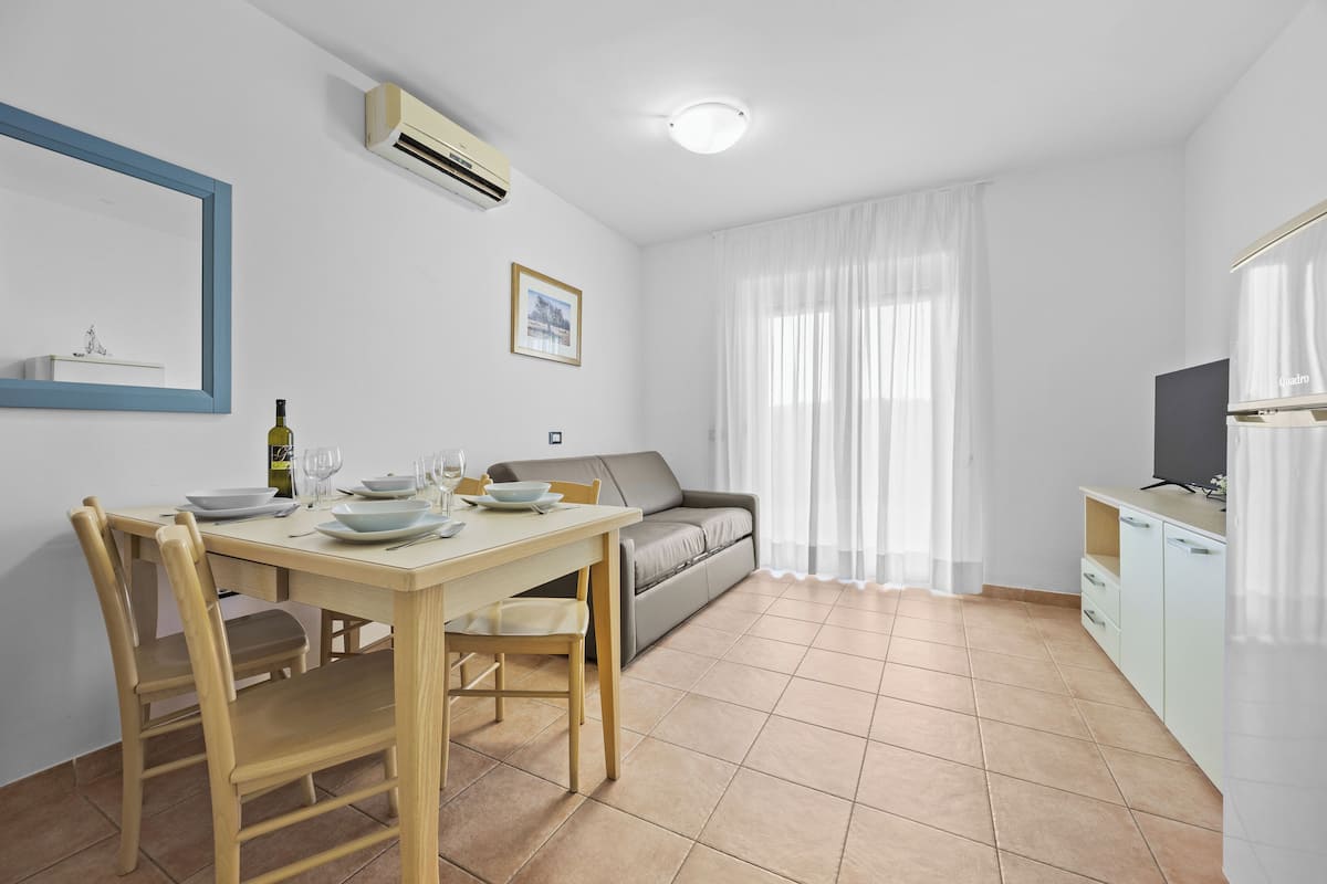 A modern, well-lit room with a dining table set for four, a sofa, a television, and a window with sheer curtains.