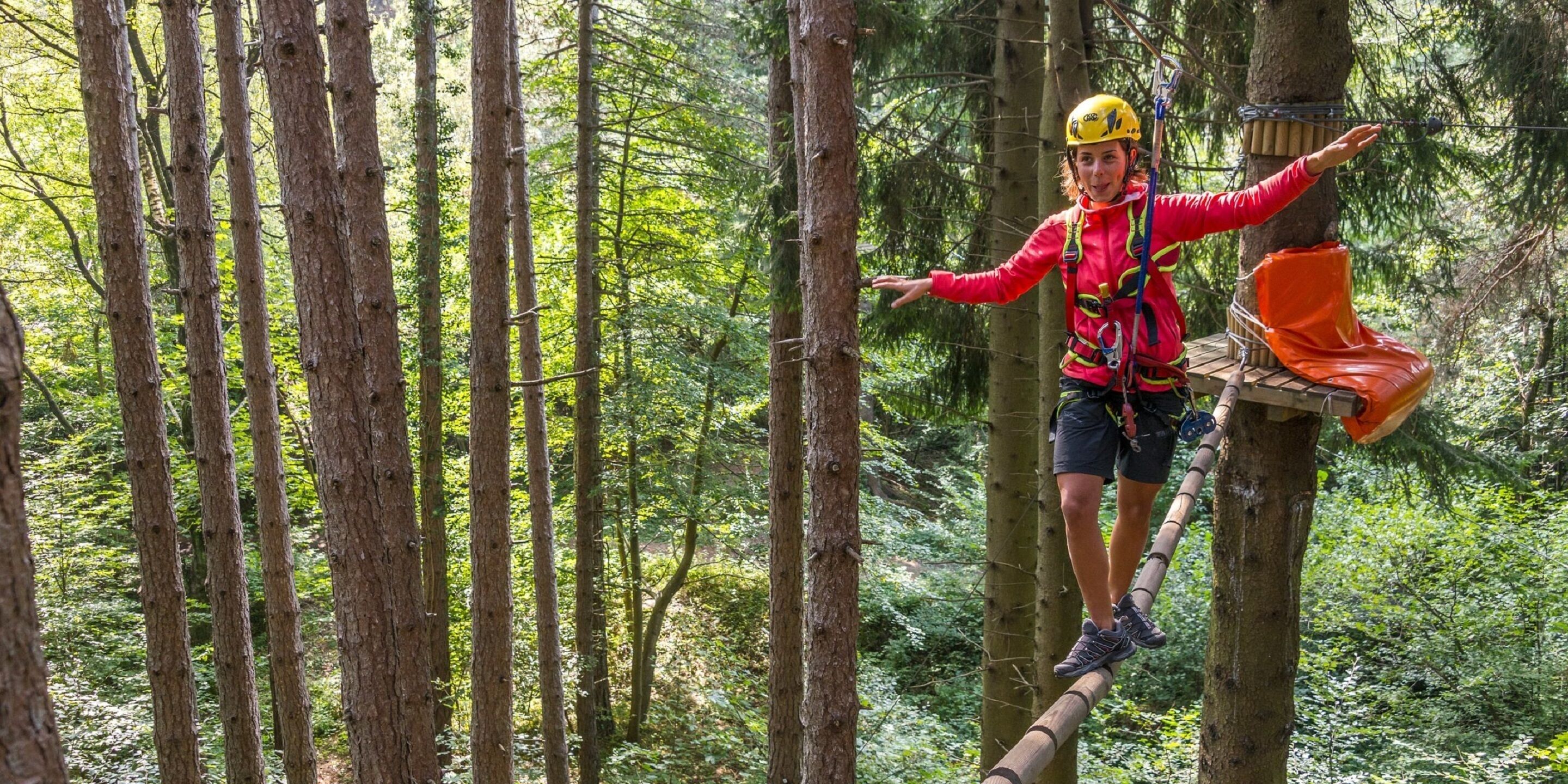 A person in a red jacket and yellow helmet navigating a forested area with a rope and harness.