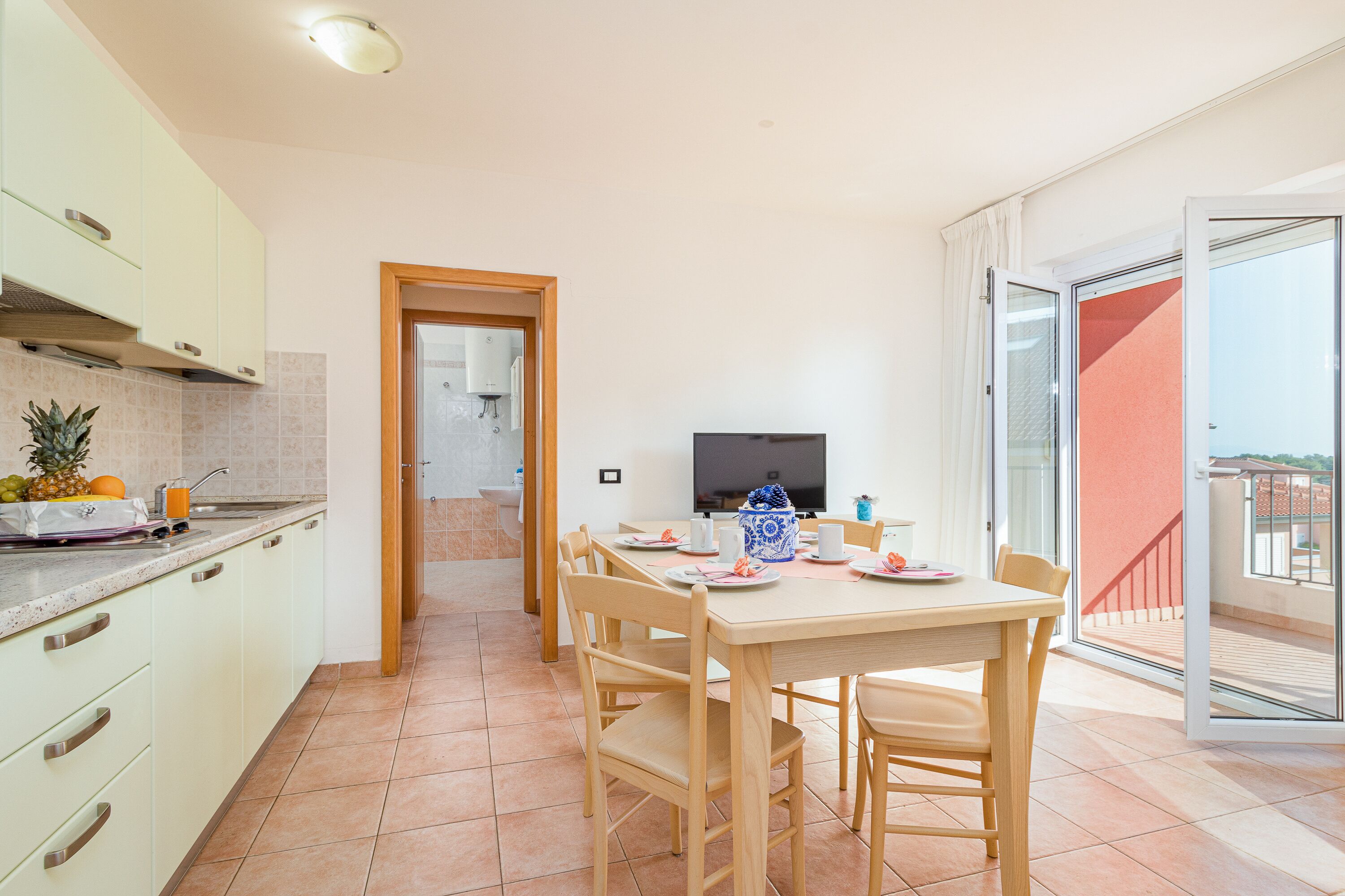 A modern kitchen with a dining area, a TV, and a balcony with a red wall.