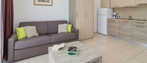 A modern living room with a sofa, a coffee table with a book and a decorative item, a kitchen with cabinets and appliances, and a framed picture on the wall.