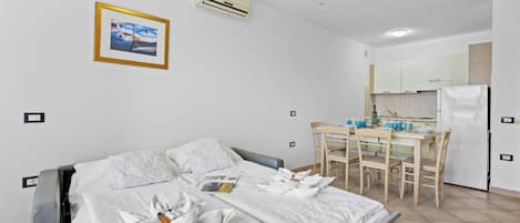 A hotel room with a bed, a dining table with chairs, a kitchen area, and an air conditioning unit.