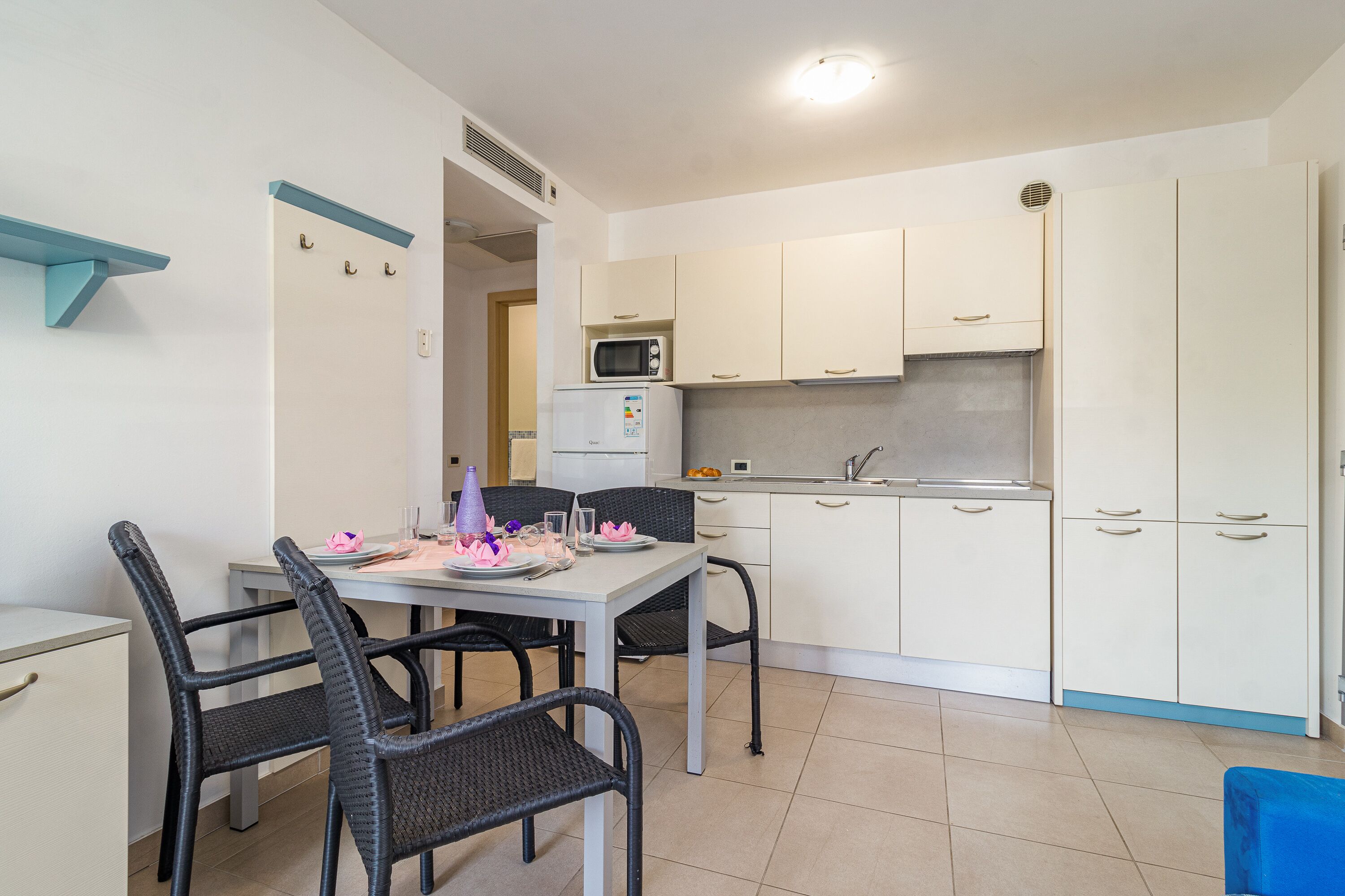 Cocina compacta con armarios blancos, microondas y un refrigerador pequeño. Hay una mesa de comedor con sillas y una silla azul.
