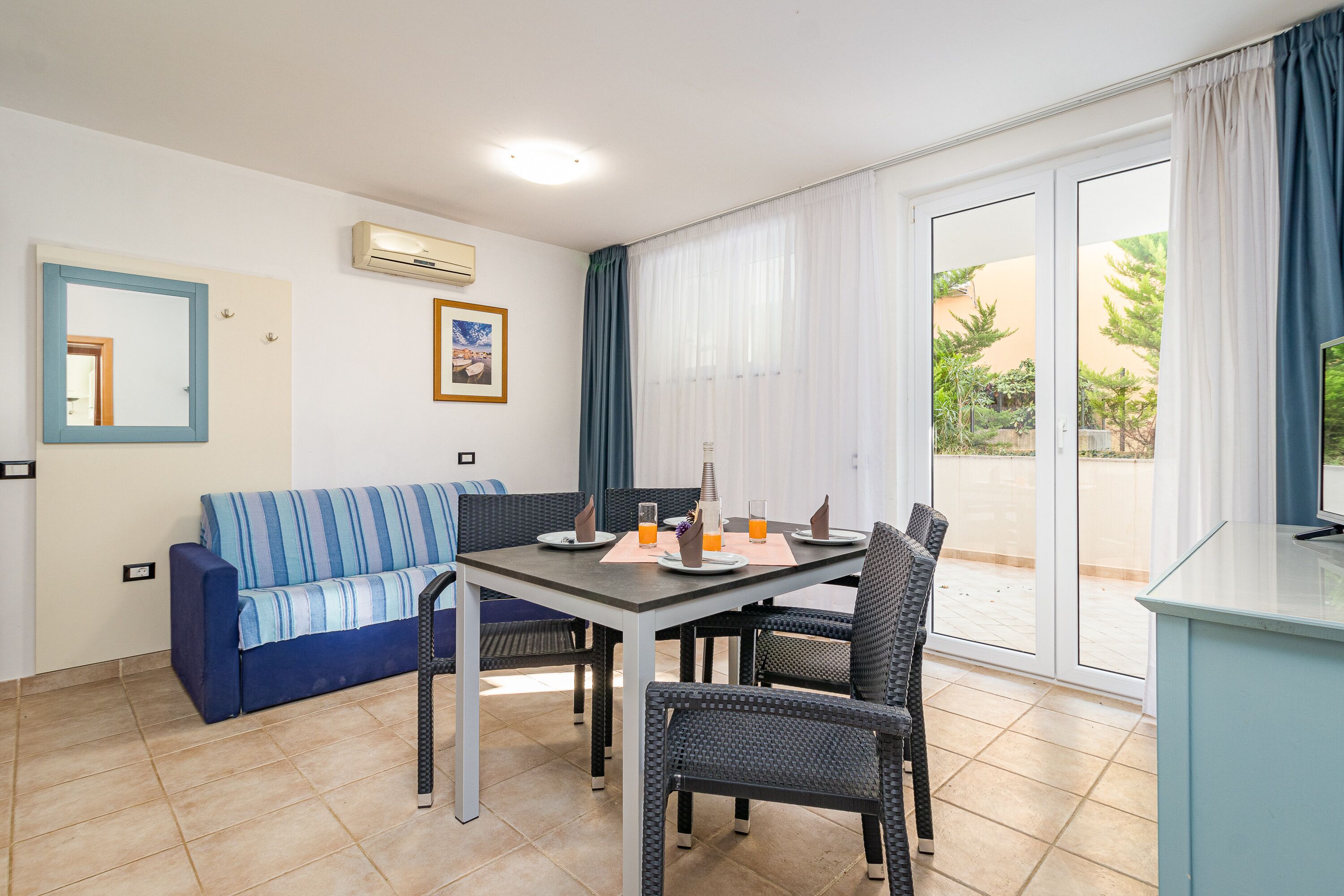 A dining area with a table set for four, a sofa, wicker chairs, and a view through a glass door to an outdoor area.