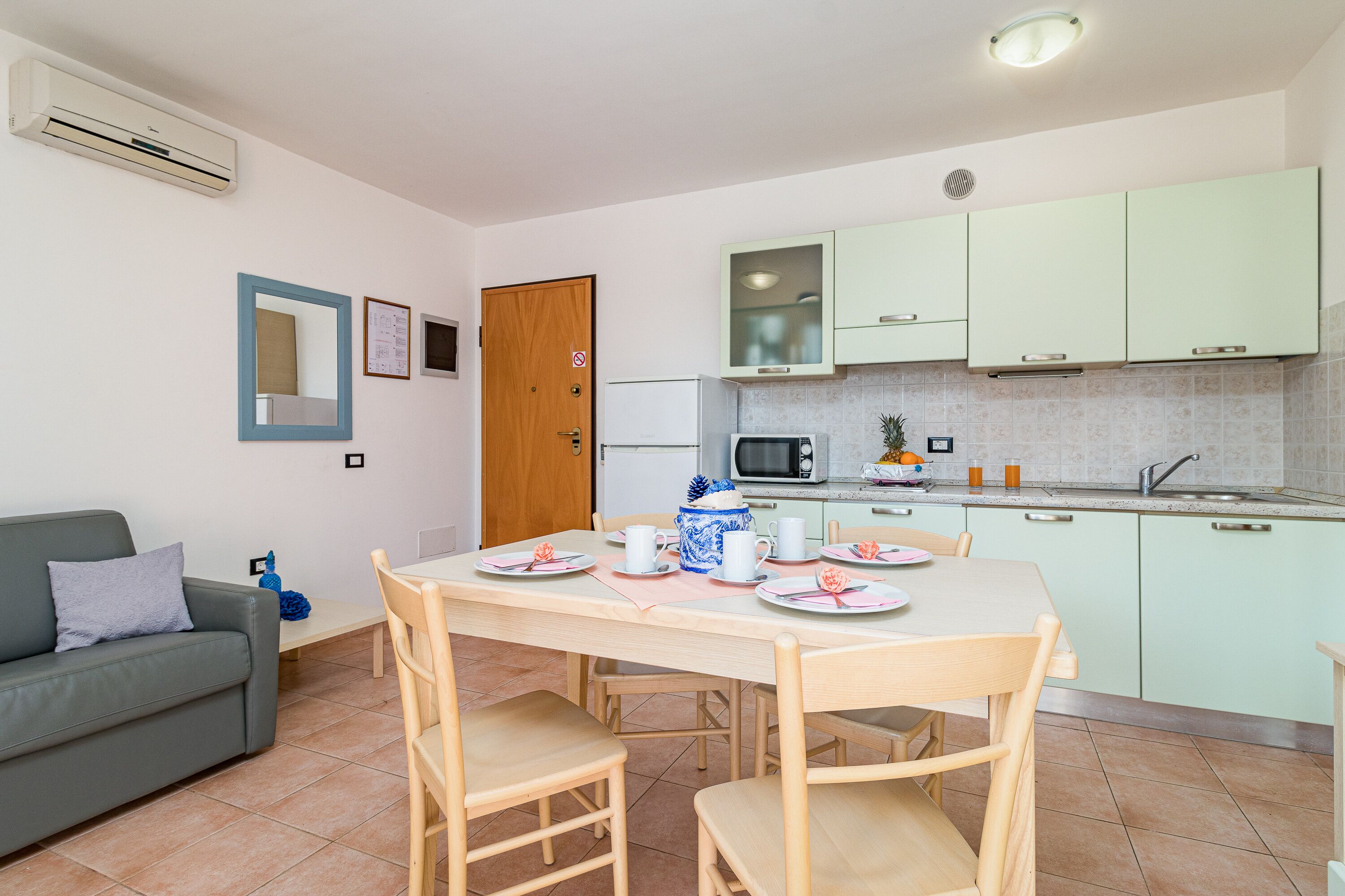 A compact kitchen with a microwave, sink, and light green cabinets. There is a dining table with chairs and a gray sofa in the living area.