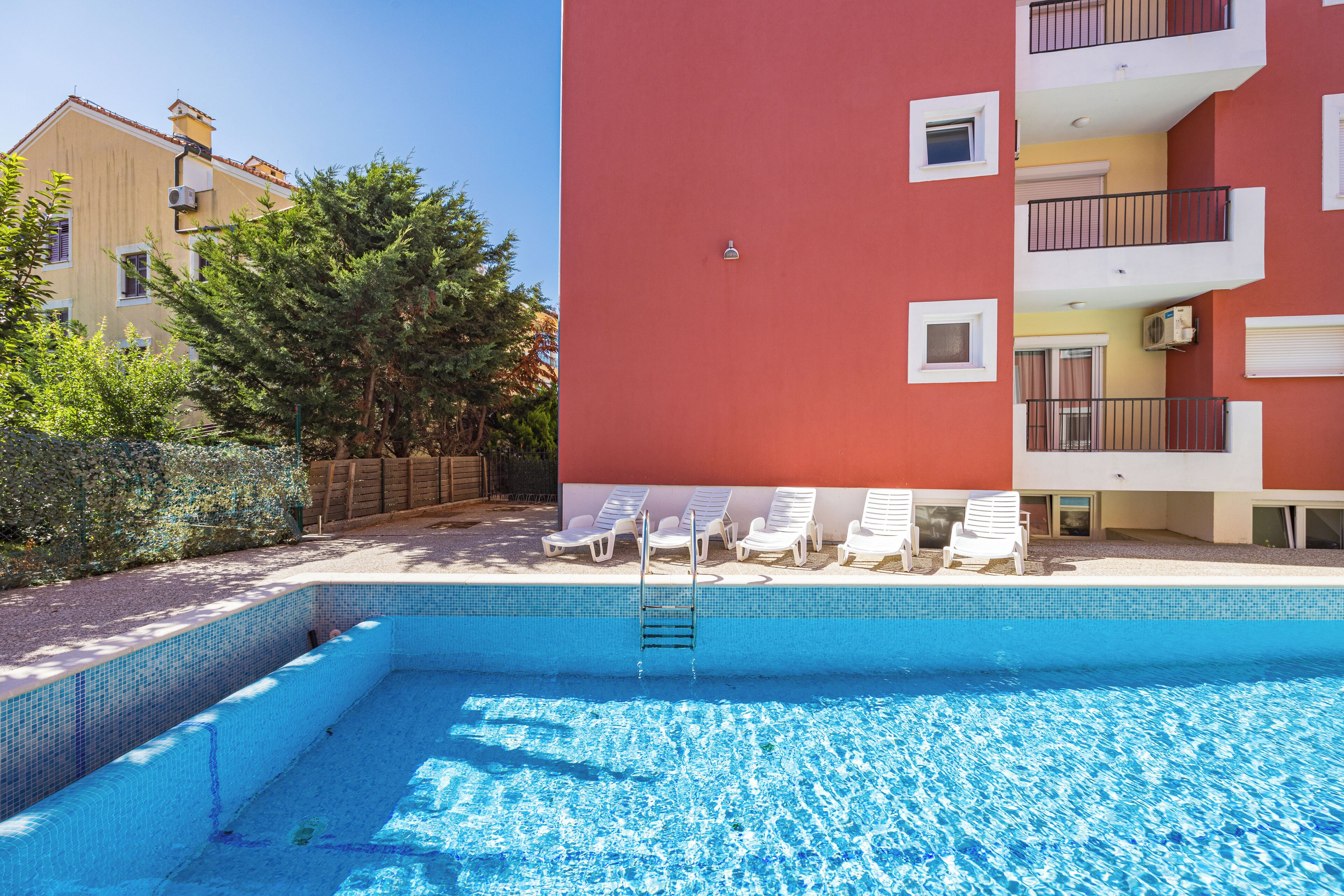 A swimming pool with a ladder, surrounded by white lounge chairs and a red building.