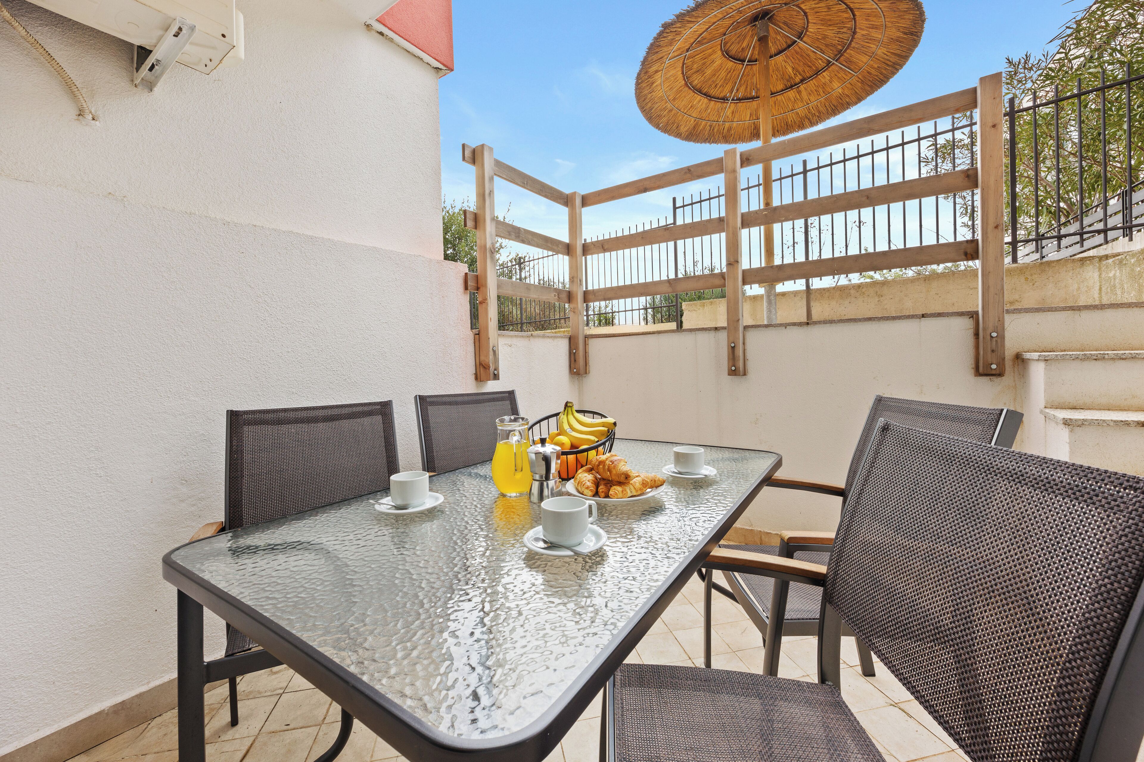 An outdoor patio with a glass table, wicker chairs, a basket of croissants, a bottle of orange juice, and a straw umbrella.