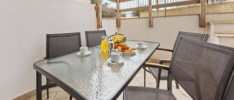 An outdoor patio with a glass table, wicker chairs, a basket of croissants, a bottle of orange juice, and a straw umbrella.