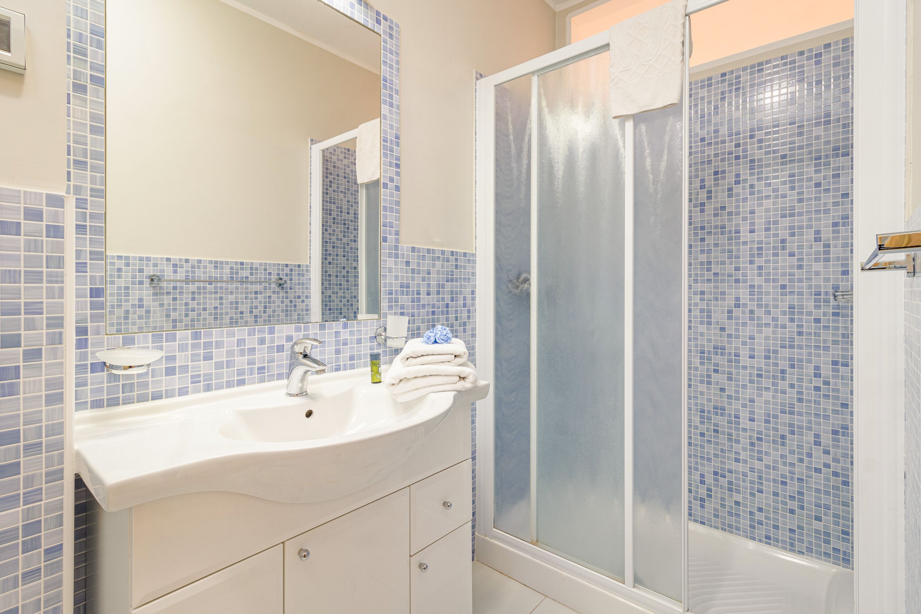 Un baño con lavamanos blanco, espejo y ducha con mosaicos azules.