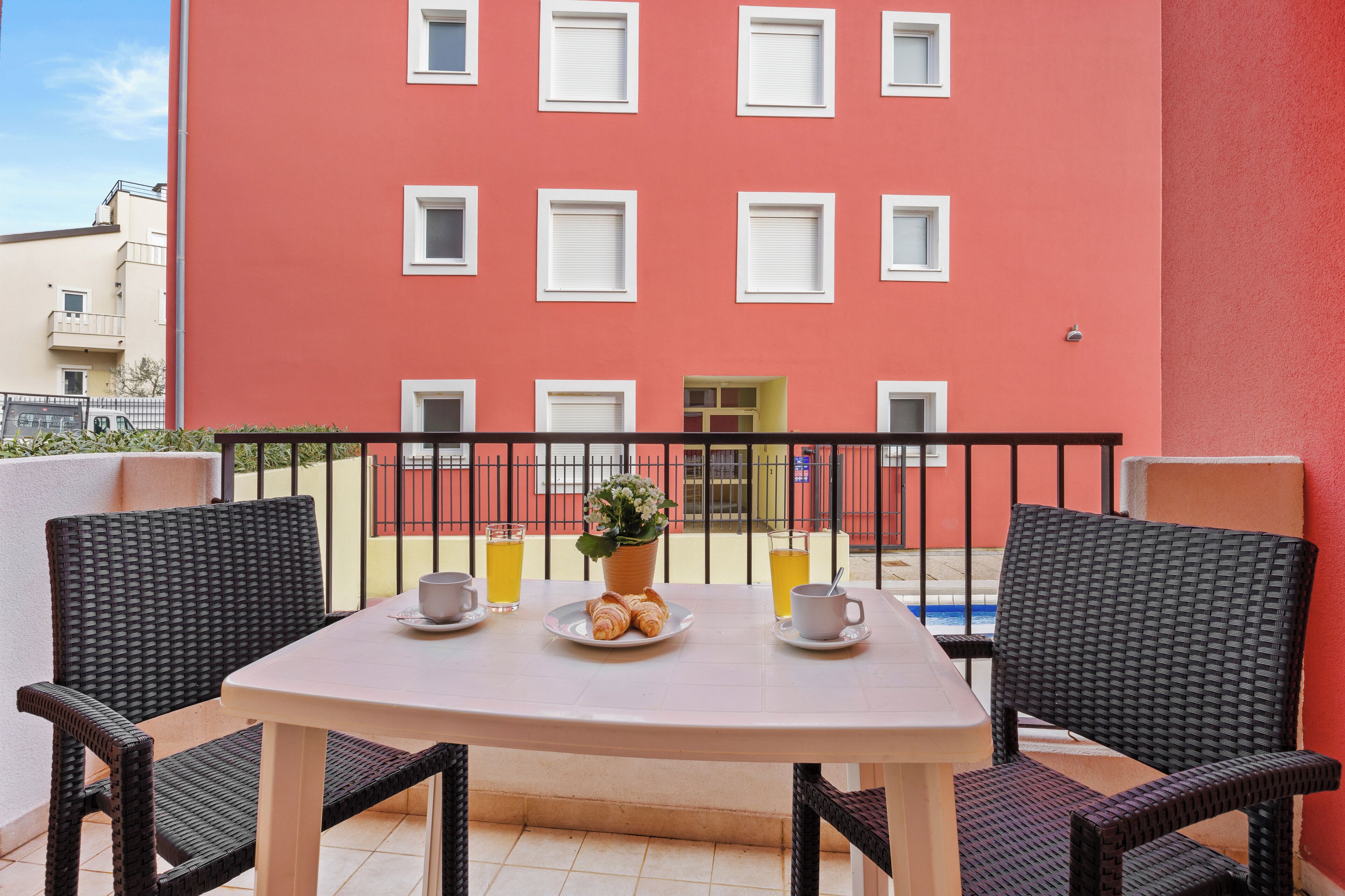 Un balcon avec une table préparée pour le petit-déjeuner, comprenant des croissants et des boissons, et un bâtiment rouge en arrière-plan.