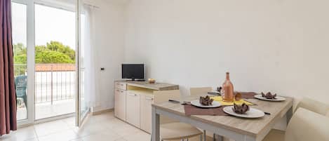 A dining area with a table set for two, a television on a stand, and a sliding glass door leading to an outdoor area.
