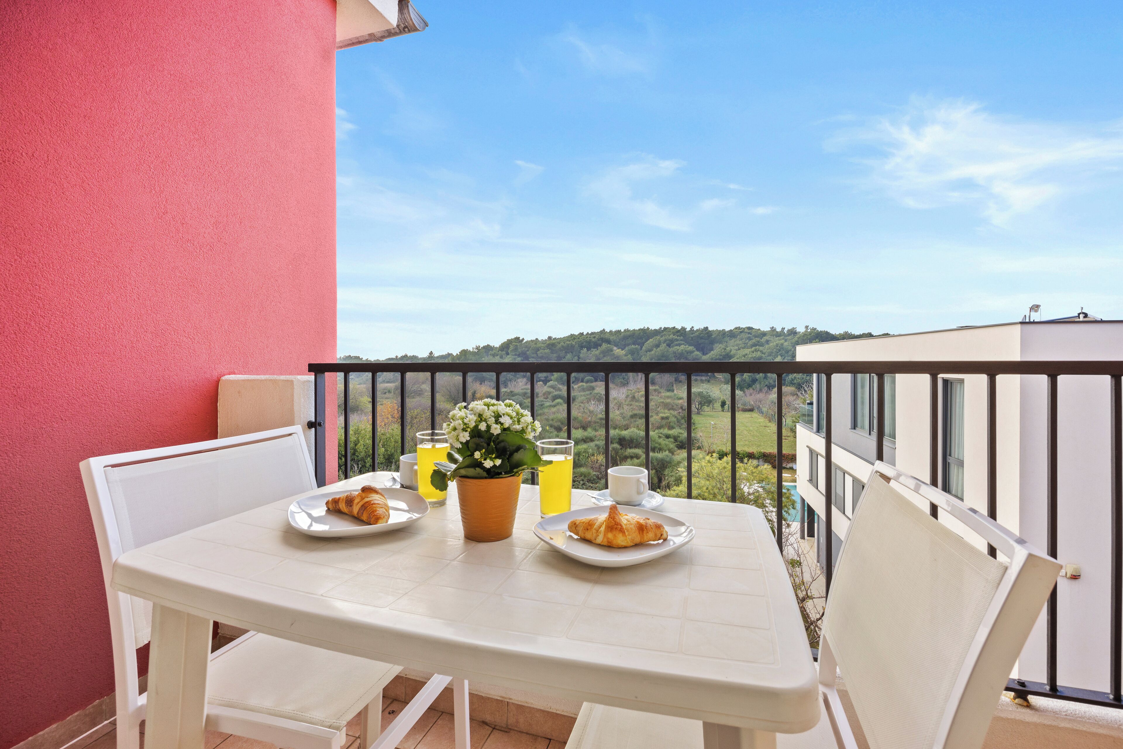 A balcony with a table set for breakfast, including croissants, a coffee cup, and a potted plant.