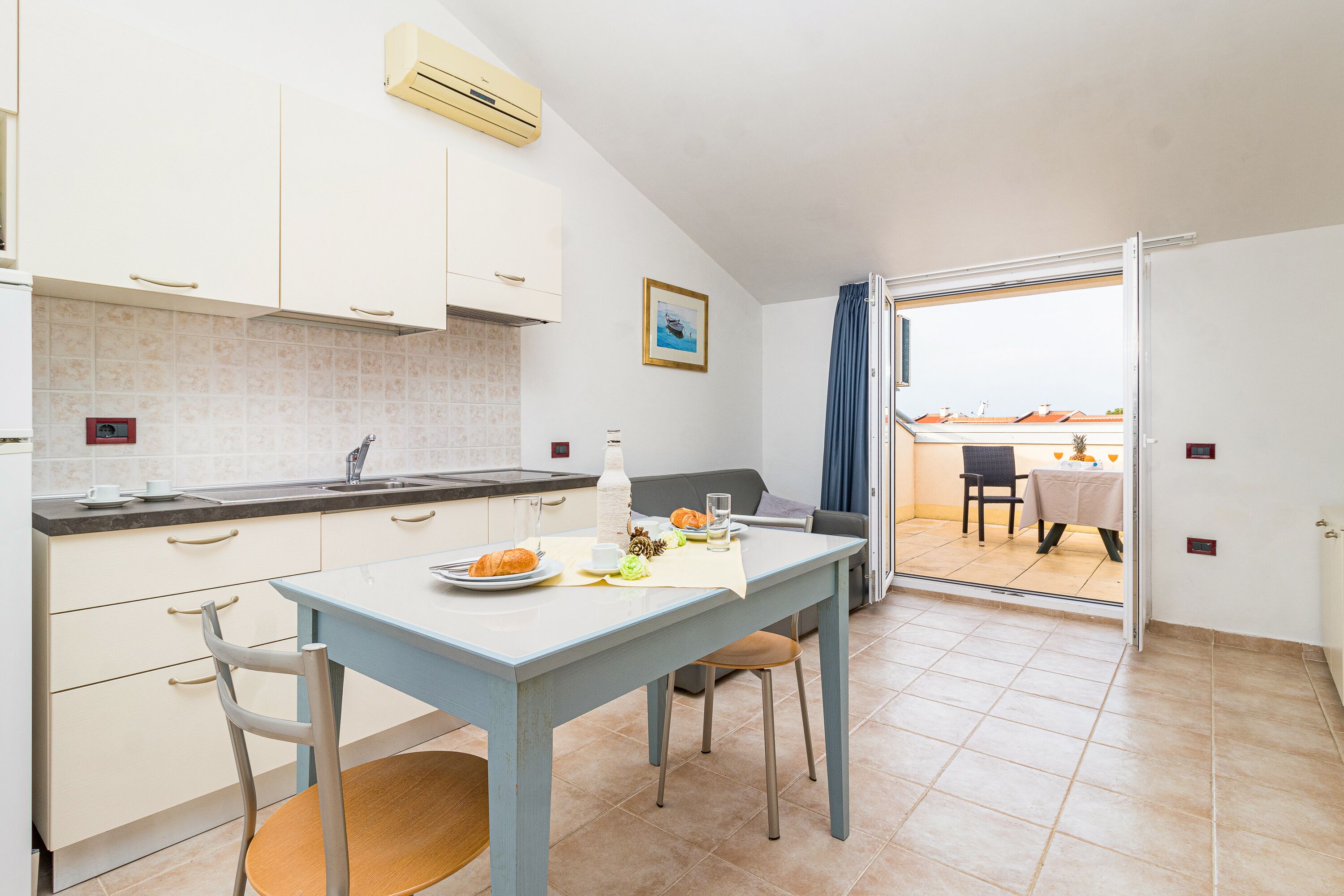 Une cuisine compacte avec un coin repas, une vue sur un espace extérieur aménagé en salon grâce à une porte vitrée, et une petite table à manger pour deux personnes.