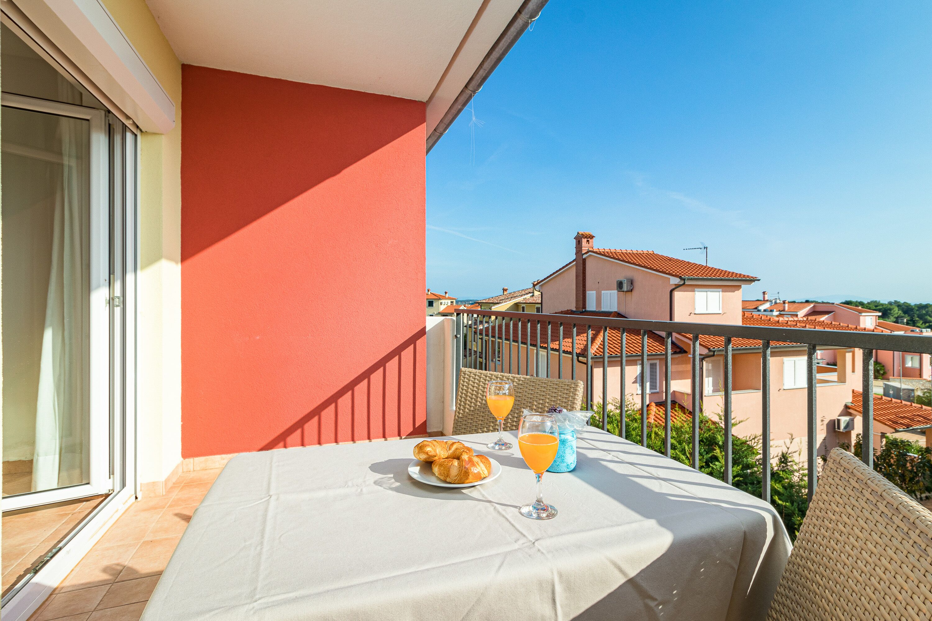 A balcony with a table set for breakfast, including croissants and orange juice, overlooking a residential area.