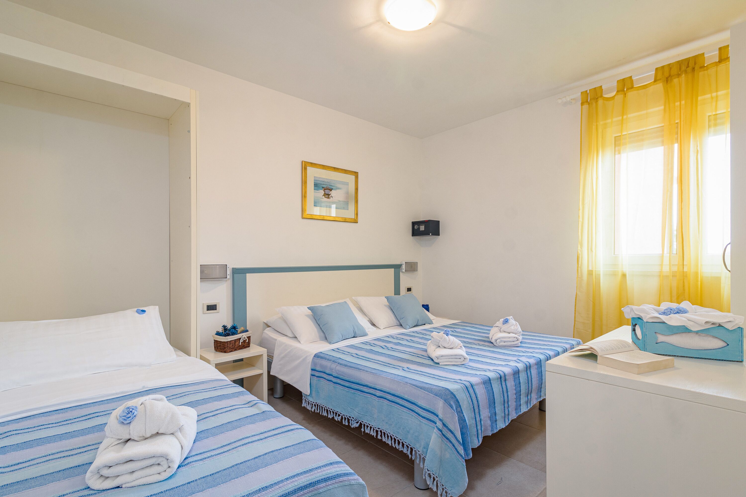 A hotel room with two beds, a bedside table, a nightstand, a framed picture on the wall, and a window with yellow curtains.