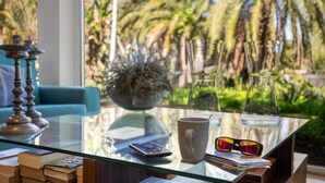 A glass table with a smartphone, a mug, sunglasses, and books, with a view of palm trees and a blue couch in the background. - Finca El Dragonal with Mountain View, Garden & Wi-Fi (Las Meleguinas, Santa Brígida)