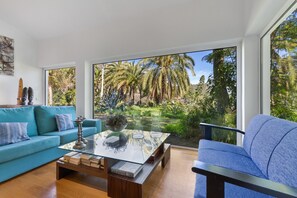 Una sala de estar moderna con una mesa de centro de vidrio, un sofá azul y vistas a un jardín tropical a través de ventanas grandes.