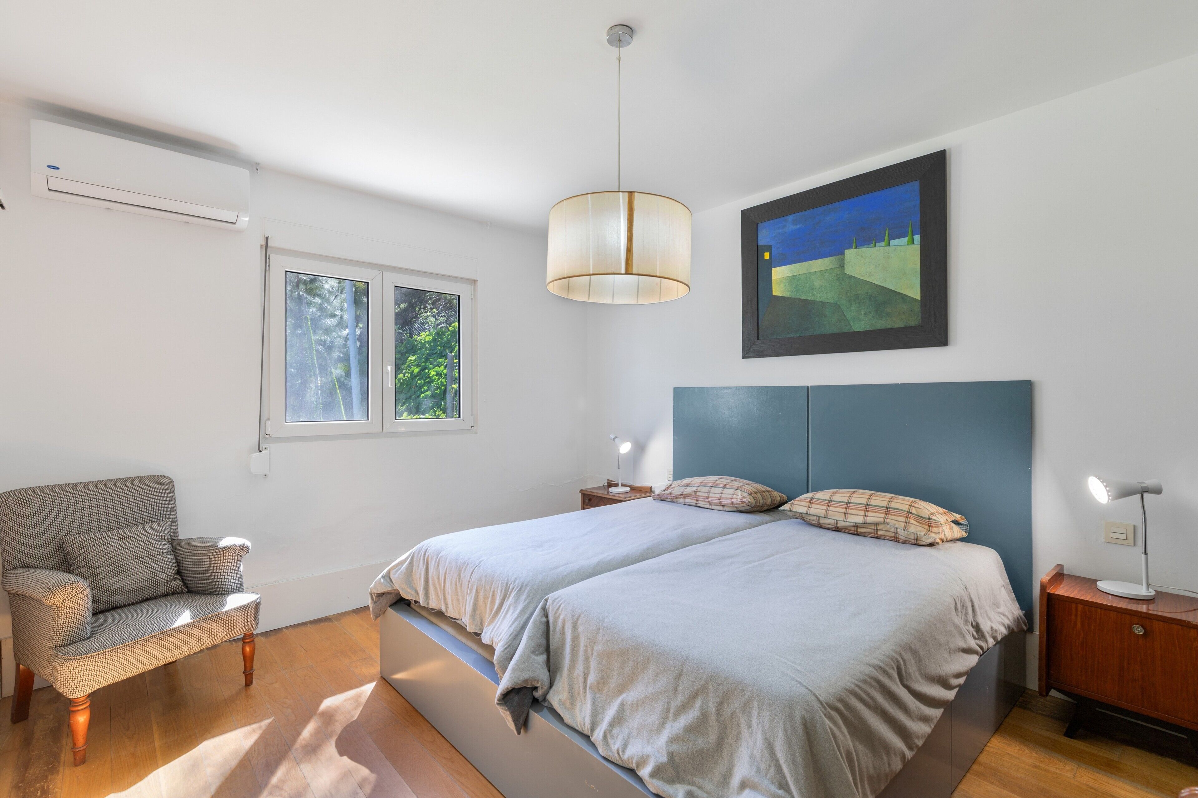Un dormitorio con cama, mesitas de noche, una silla, una ventana y un cuadro.