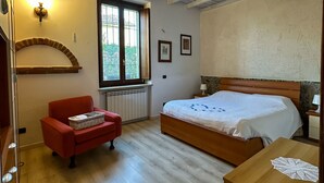 A bedroom with a wooden bed, a red armchair, a wooden dresser, and a window with a view of greenery. - Appartamento in storica Cascina Lombarda Cusago (Cascina Cusago di Sotto)