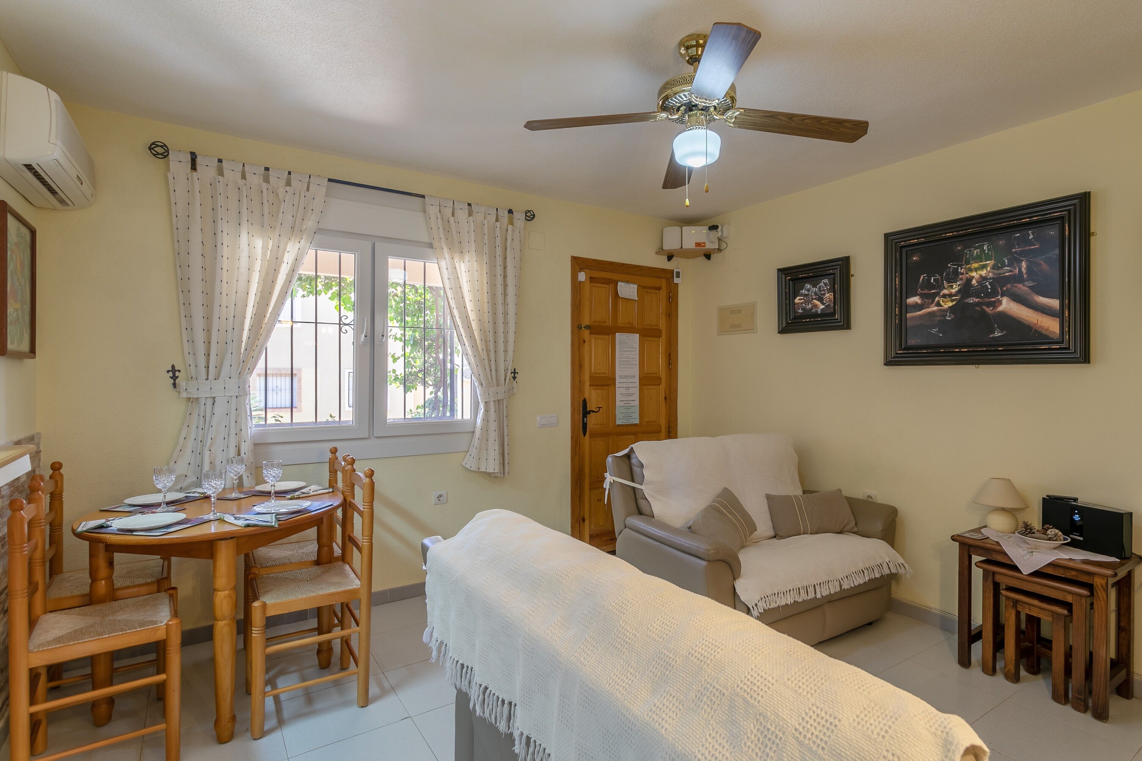 Una habitación con una mesa de comedor preparada para cuatro personas, un sofá, un ventilador de techo y una pequeña mesa de madera con una lámpara.