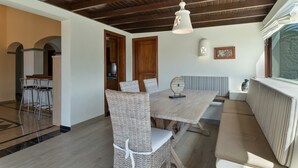 Un coin repas avec une table en bois, des chaises en osier, un banc et un plafond avec des poutres apparentes.
