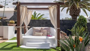 Un espace extérieur aménagé avec des sièges blancs, un pergolas en bois, des coussins et un mur en pierre en arrière-plan.