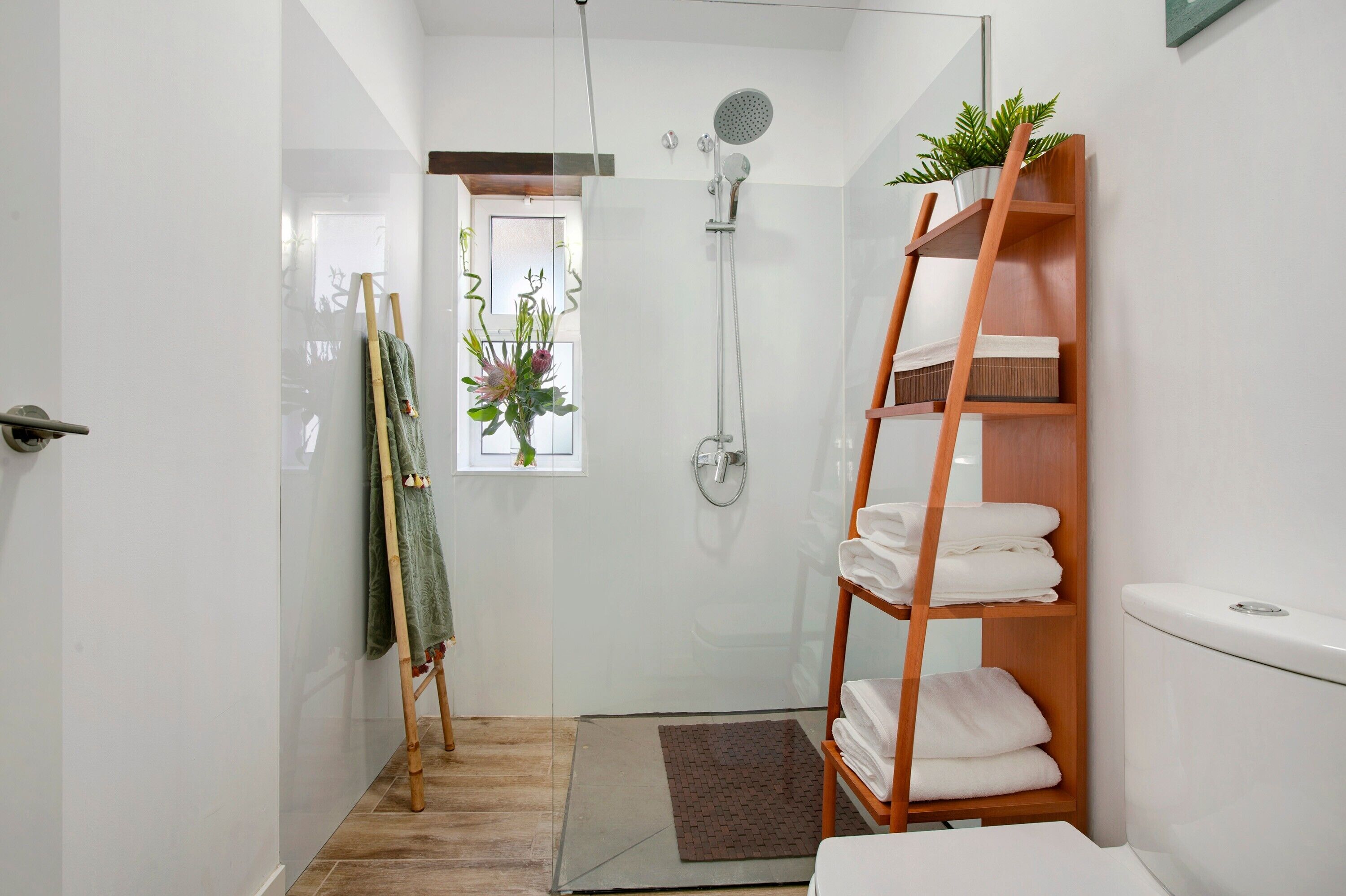Un baño moderno con cabina de ducha de vidrio, un perchero de madera, un inodoro y una pequeña ventana con una planta.
