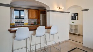 A kitchen with a refrigerator, wooden cabinets, and a window with curtains. There are three white bar stools and a white shelf. - Villa Los Alisios 58 with Mountain Views, Pool, Wi-Fi, Terraces and Garden (Playa Blanca)