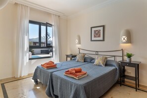 A double bed with a headboard, two bedside tables, a window with curtains, a framed picture on the wall, and a small plant on a bedside table.