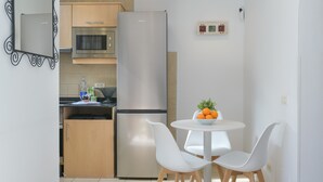 Una cocina compacta con refrigerador, microondas y gabinetes de madera. Hay una mesita con un tazón de fruta y dos sillas blancas.
