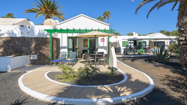 Un edificio con fachada blanca, detalles en verde y un área de estar al aire libre cubierta con mesas y sillas.