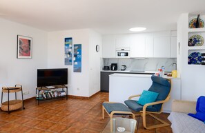 A modern living room with a flat-screen TV, a bookshelf, a dining area with a kitchen in the background, and a small table with a chair.