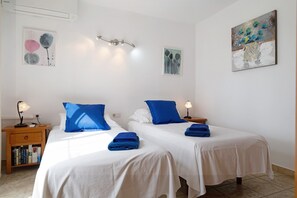 Ein Hotelzimmer mit zwei Betten, weißer Bettwäsche, blauen Kissen, einem Nachttisch mit Lampe und Bildern an den Wänden.