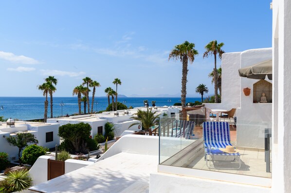 A balcony with a clear view of the sea, palm trees, and a white building with a glass railing.